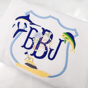 May include: White fabric with an embroidered design featuring a blue shield shape. Inside the shield are the letters "RBJ" in blue, with a sailboat, fish, and a hummingbird. The text "Ruby Dear Designs" is also present.