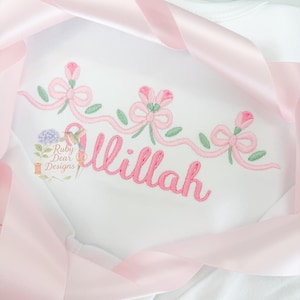 Puede incluir: Body blanco con bordado rosa que representa flores, lazos y el nombre "Illah" en escritura cursiva rosa. Una cinta de raso rosa está drapeada alrededor de la prenda. Las hojas verdes completan el diseño.
