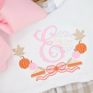 Puede incluir: Un pelele blanco con una letra E bordada en rosa, calabazas, hojas y trozos de pastel. El texto "Ruby Dear Boutique" está bordado en rosa.