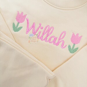 May include: Cream-colored sweatshirt with the name "Willah" embroidered in pink script. Pink tulip flowers with green stems and leaves flank the name. The sweatshirt has a scalloped edge.