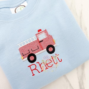 May include: Light blue baby shirt with a red embroidered fire truck design. The fire truck has black wheels and a white ladder. The name "Rhett" is embroidered in red script below the truck.