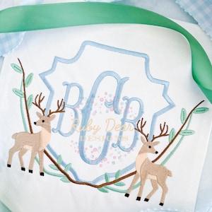 May include: A blue and white monogram with the letters "RDB" in a decorative frame. Two brown deer are on either side of the monogram, surrounded by green leaves.