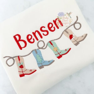 May include: Cream-colored fabric with embroidered cowboy boots in red, blue, and tan, connected by a rope. The name "Benser" is embroidered in red above the boots. A floral and hummingbird design is in the upper right corner.