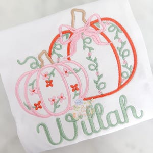 May include: A white shirt featuring an embroidered design of two pumpkins. One pumpkin is pink with small red and pink flowers, while the other is orange with green vines. A pink bow sits atop the orange pumpkin. The name Willah is embroidered in green.