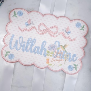 May include: Embroidered name plaque with a scalloped edge, featuring the name "Willah" in light blue script. Adorned with pink bows, butterflies, and floral accents. Includes white ribbons for hanging. The plaque is a soft pink and white color.