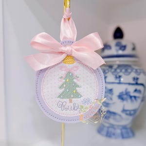 Puede incluir: Adorno navideño redondo con un diseño bordado de un árbol de Navidad verde, lazos rosas y el nombre "Ruby". Tiene un borde lavanda y un lazo de cinta de raso rosa. Una pinza dorada sujeta el adorno.