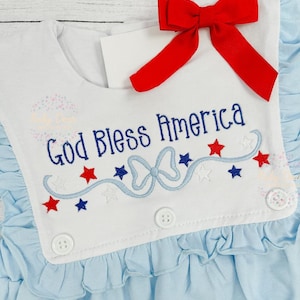 May include: A white baby dress with blue ruffles and a red bow. The dress has an embroidered design that says "God Bless America" with stars and a blue ribbon.