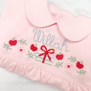 Pode incluir: Roupa de bebé rosa com gola Peter Pan. A frente apresenta bordados de maçãs vermelhas, flores, livros e o nome "Willah" em fio azul claro. Um laço vermelho também é bordado.