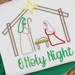 May include: White t-shirt with an embroidered Nativity scene. The design features a star, stable, and figures of Joseph, Mary, and baby Jesus. The words "O Holy Night" are embroidered in green script below the scene.