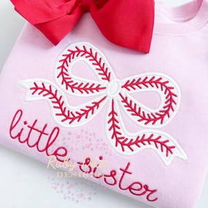 May include: Pink sweatshirt with a white and red baseball-stitched bow and the text "little sister" embroidered on the front.