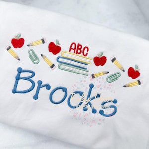 May include: White fabric embroidered with school-themed designs. Features red apples, yellow pencils, green paperclips, and a stack of books with "ABC" in red. The name "Brocks" is embroidered in blue.