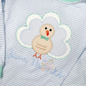 Könnte beinhalten: Ein hellblau-weiß gestreiftes Langarmshirt mit einem Truthahn-Applikation. Der Truthahn ist beige mit einer grünen Fliege und einem orangefarbenen Schnabel. Das Shirt hat den gestickten Text "Give Thanks".
