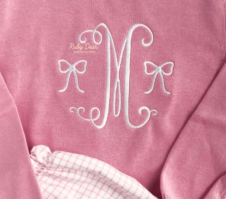 May include: Pink knitted sweater with a white embroidered letter M and two bows. The sweater is folded over a pink and white gingham fabric.