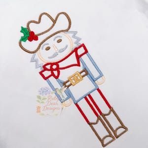 Könnte beinhalten: Weißes T-Shirt mit einem gestickten Cowboy-Nussknacker-Design. Der Nussknacker hat ein weißes Gesicht, einen braunen Hut mit Stechpalme und eine rote Fliege. Das Design umfasst blaue und rote Akzente sowie braune Stiefel.