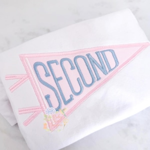 May include: A white shirt featuring an embroidered pink pennant flag design with the word SECOND stitched in light blue letters. The pennant includes two pink ties on the left side and a small floral graphic at the bottom corner of the flag.