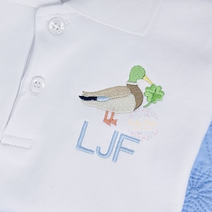 May include: White polo shirt with an embroidered duck holding a four-leaf clover. The shirt has a blue collar and the letters "LJF" are embroidered below the duck.