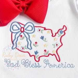 Può includere: Un body bianco per neonato con un ricamo rosso, bianco e blu degli Stati Uniti con fiori e stelle. Il testo "God Bless America" è ricamato sotto il disegno.