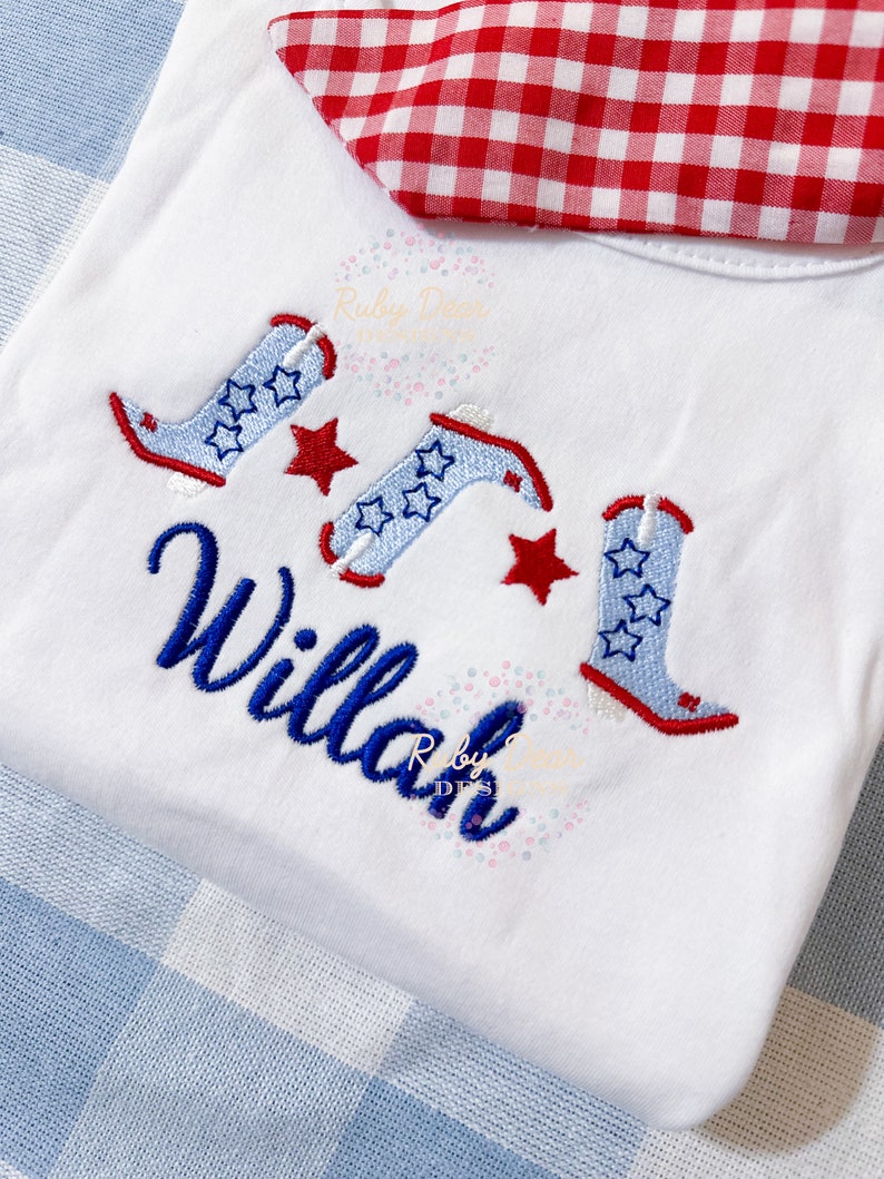 May include: White onesie with a red and white checkered fabric on the top. The onesie has blue and white cowboy boots with red stars embroidered on it. The name "Willa" is embroidered in blue cursive lettering.