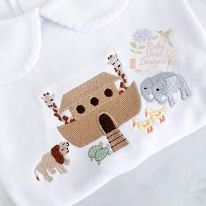 May include: White collared shirt with an embroidered Noah's Ark scene. The design includes a brown ark, giraffes, an elephant, ducks, a lion, and a turtle. The shirt features a scalloped collar and the text "Ruby Dear Designs".