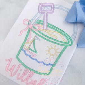 Puede incluir: Camiseta blanca bordada con un diseño de cubo de playa, un velero, un sol y el nombre "Willah" en escritura rosa. El cubo tiene un asa y una pala. Un lazo azul es visible en el fondo.