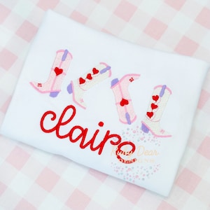 May include: White t-shirt featuring four embroidered cowboy boots in pink, white, and lavender, each adorned with red heart accents. The name "claire" is embroidered in red script. A cute design for Valentine's Day.