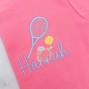 May include: A bright pink t-shirt featuring an embroidered design of a light blue tennis racket, a yellow tennis ball, and the name Hannah in light blue script lettering. A small floral and hummingbird graphic is positioned below the tennis ball.