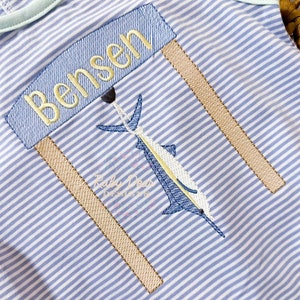 May include: A blue and white striped onesie with a brown and yellow sign that says "Bensen" above a blue marlin fish.
