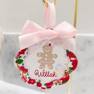 May include: A decorative ornament featuring a gingerbread person design. The ornament has a pink bow and a floral patterned border with the name "Willah" embroidered on it. The gingerbread person has a pink bow and skirt.