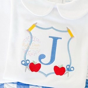 May include: White collared shirt with a blue and white embroidered shield featuring a blue letter J, two red apples, two yellow pencils, and two silver scissors.