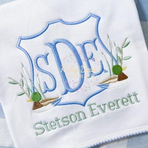 May include: A white baby blanket with blue trim and a blue and green embroidered monogram with the initials "SDE" and the name "Stetson Everett". The monogram is surrounded by two embroidered ducks and green leaves.