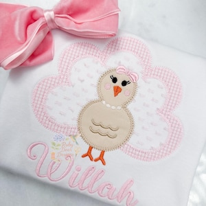 Puede incluir: Una camiseta blanca con un diseño de nube rosa a cuadros y un pollito de dibujos animados con un lazo. La camiseta tiene el nombre "Willah" bordado en rosa. Un lazo de terciopelo rosa está colocado en la parte superior izquierda.