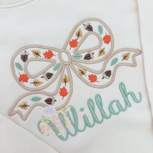 May include: Cream sweatshirt with an embroidered bow design filled with autumn leaves and acorns in orange, brown, and green. The name "Willah" is embroidered in a cursive, light green font below the bow.