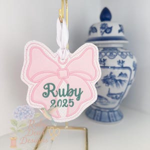 Op de afbeelding: Een roze strikvormige ornament met de naam "Ruby" en het jaar "2025" in groen geborduurd. Het ornament hangt aan een goudkleurige standaard met een wit lint. Een blauw-witte decoratieve pot is op de achtergrond te zien.