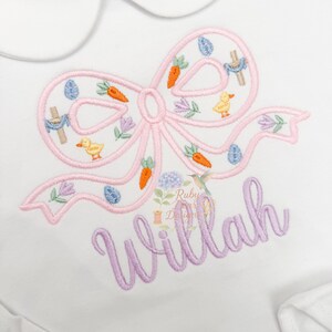 May include: White baby onesie with a Peter Pan collar. The onesie features a pink embroidered bow with Easter-themed details, including carrots, eggs, and a duckling. The name "Willah" is embroidered in lavender script below the bow.