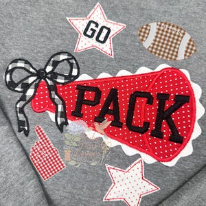 Puede incluir: Una sudadera gris con parches bordados. Los parches incluyen una forma de megáfono rojo con la palabra "PACK", un balón de fútbol, estrellas y un lazo. Los parches son rojos, blancos y negros.