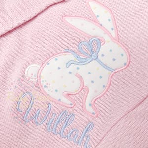 以下が含まれることがあります： 青いリボンが付いた白いウサギのアップリケが付いたピンクのニットセーター。ウサギは水玉模様です。セーターは「Willah」という名前が青い刺繍でパーソナライズされています。