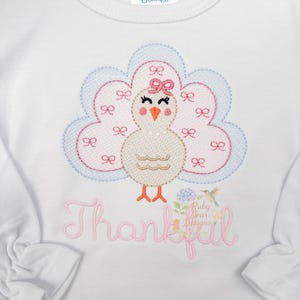May include: White long-sleeved shirt featuring an embroidered turkey design. The turkey has a pink bow, and is surrounded by a blue and pink cloud-like shape. The word "Thankful" is embroidered in pink script below the turkey.
