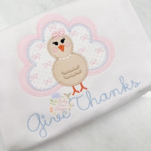 Może przedstawiać: Biała tkanina z haftowanym wzorem indyka. Indyk jest brązowy z różowym i niebieskim ogonem w kształcie chmury. Tekst "Give Thanks" jest wyszyty na niebiesko poniżej. Widoczne jest małe logo.