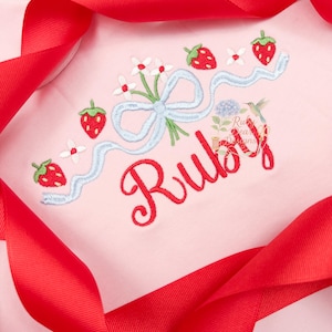 May include: Pink baby onesie with embroidered strawberries, flowers, and a blue ribbon design. The name "Ruby" is embroidered in red script. A red ribbon frames the onesie.