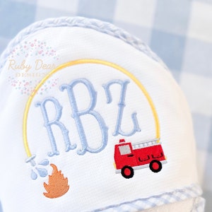 May include: A white hooded towel with a blue and white gingham border. The towel has a yellow and blue embroidered fire truck with a hose and a fire. The towel also has a blue embroidered monogram "RBZ" in a yellow circle.