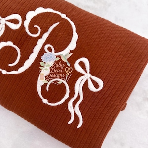 May include: A rust-colored fabric with white embroidered details. The design includes a large letter "R" with floral and hummingbird accents, and a bow. The text "Ruby Dear Designs" is also embroidered on the fabric.