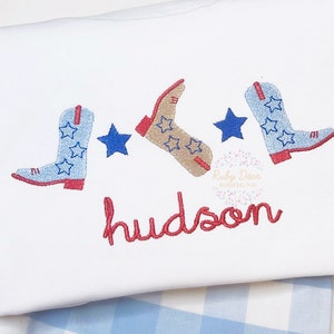 Puede incluir: Una camiseta blanca con botas de vaquero y estrellas bordadas en rojo, azul y marrón. El nombre "hudson" está bordado en rojo debajo de las botas.