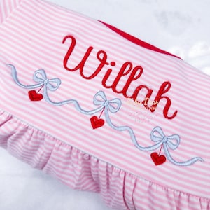 May include: Pink and white striped baby dress with a red embroidered name "Willah" and a design of silver bows and red hearts.