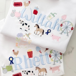 Może przedstawiać: Dwie białe koszulki z haftowanymi wzorami. Jedna koszulka ma imię "Willah" w jasnoniebieskim skrypcie, otoczone haftowanymi obrazami krowy, konia i diabelskiego młyna. Druga koszulka mówi "Rhett" z podobnymi wzorami.