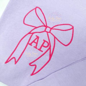 Op de afbeelding: Een paars sweatshirt met een roze geborduurde strik en de letters "AP" in de strik.