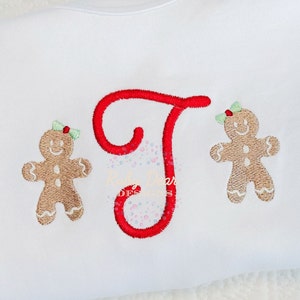 May include: A white onesie with a red embroidered letter "T" and two gingerbread people on either side. The gingerbread people are brown with white icing and have green bows on their heads.