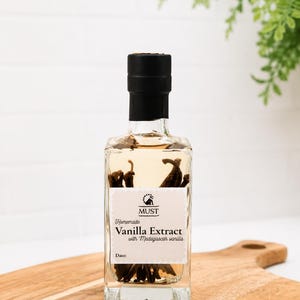 Könnte beinhalten: Eine klare Glasflasche gefüllt mit bernsteinfarbener Flüssigkeit und dunklen Vanilleschoten. Das weiße Etikett trägt den Text MUST, Homemade Vanilla Extract with Madagascar vanilla und eine leere Datumszeile. Schwarzer Verschluss und quadratischer Boden.