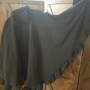 May include: A black hooded cape with a ruffled edge. The cape is made of a soft material and hangs from a hanger. The hood is visible at the top, and the ruffled edge adds a decorative touch to the garment.