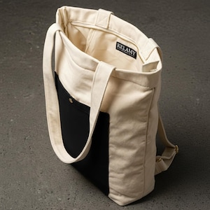 Peut inclure: Sac fourre-tout en toile de couleur crème avec une poche noire et des bretelles. Le sac a une fermeture éclair et une petite étiquette qui dit "KELAMY". Le sac est ouvert, révélant la doublure intérieure.