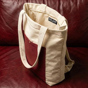 May include: Cream-colored canvas tote bag with a burgundy pocket and accents. The bag has a zipper closure and a shoulder strap. The interior is a light beige color. The brand name "KELAMY" is visible on a tag inside the bag.